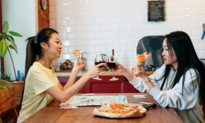 two sisters raising a glass of red wine as they play Monopoly and eat pizza in their kitchen.