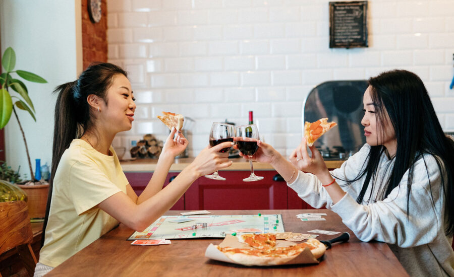 two sisters raising a glass of red wine as they play Monopoly and eat pizza in their kitchen.