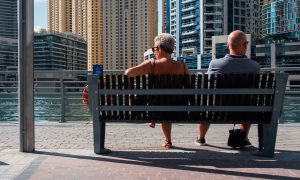 A couple in their mid-50s sits on a bench along the water in front of of condo buildings.