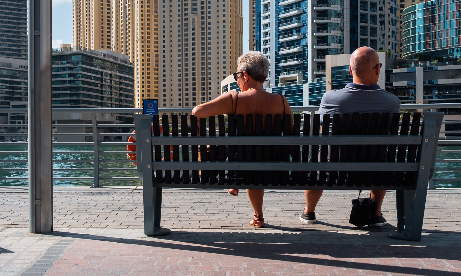 A couple in their mid-50s sits on a bench along the water in front of of condo buildings.
