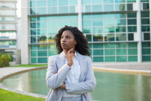 Serious pensive business leader thinking over strategy outside. Young black woman wearing formal suit, standing near office building, touching chin and looking away. Pensive businesswoman concept