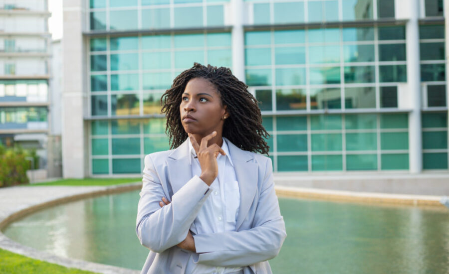 Serious pensive business leader thinking over strategy outside. Young black woman wearing formal suit, standing near office building, touching chin and looking away. Pensive businesswoman concept
