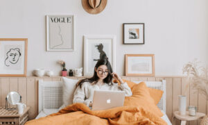 Woman in the bedroom of her apartment, surrounded by her items and in bed with her computer trying to figure things out.