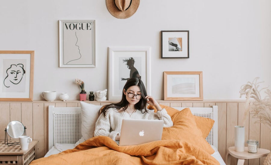 Woman in the bedroom of her apartment, surrounded by her items and in bed with her computer trying to figure things out.