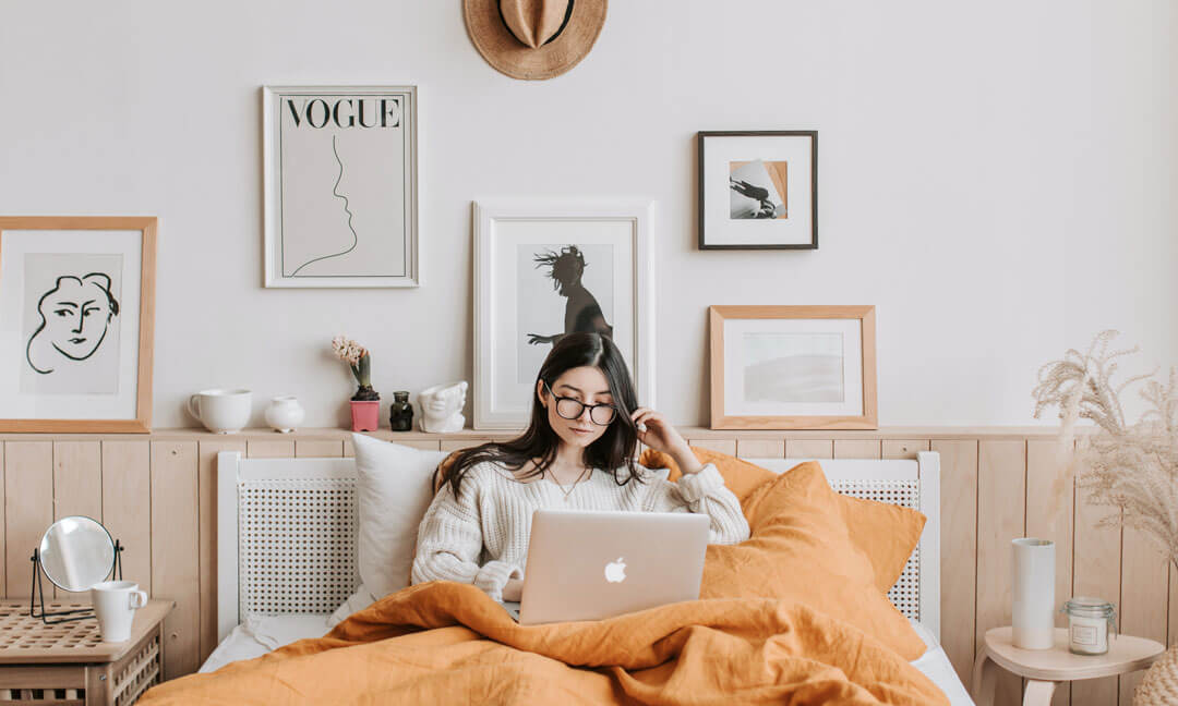 Woman in the bedroom of her apartment, surrounded by her items and in bed with her computer trying to figure things out.