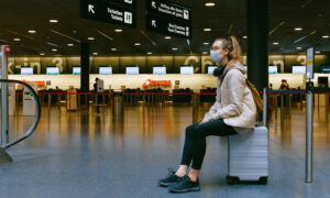 A woman wearing a mask, while sitting on a suitcase waiting for her flight gate to be announced.