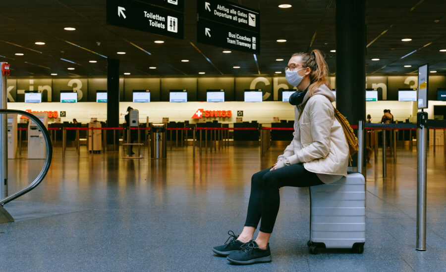 A woman wearing a mask, while sitting on a suitcase waiting for her flight gate to be announced.