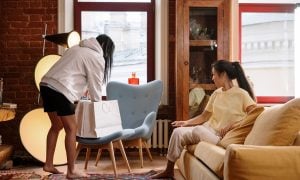 Two women in their home, as one shows the other the things she bought that day. She's reaching in the shopping bag.