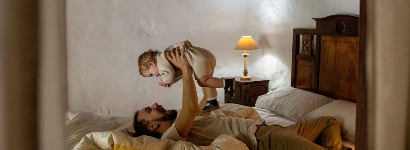 A father and a baby playing together on a bed at the cottage.