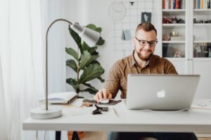 man with glasses sitting at desk and reading laptop computer screen