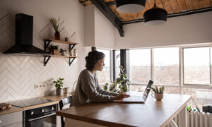 A woman in a kitchen, at the island, looking up her credit score on a computer.