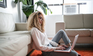 A woman is sitting on the floor of her living room, researching investing on her laptop.