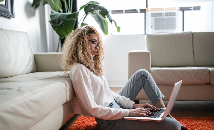 A woman is sitting on the floor of her living room, researching investing on her laptop.