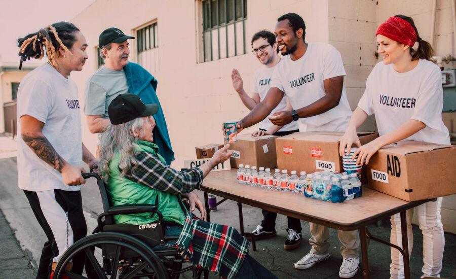 Five people are seen in a photo giving food to a man in a wheel chair