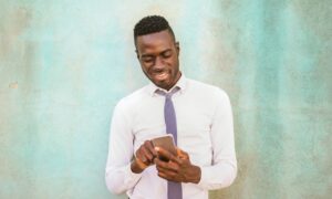 A smiling man looking at his phone while leaning against a wall.