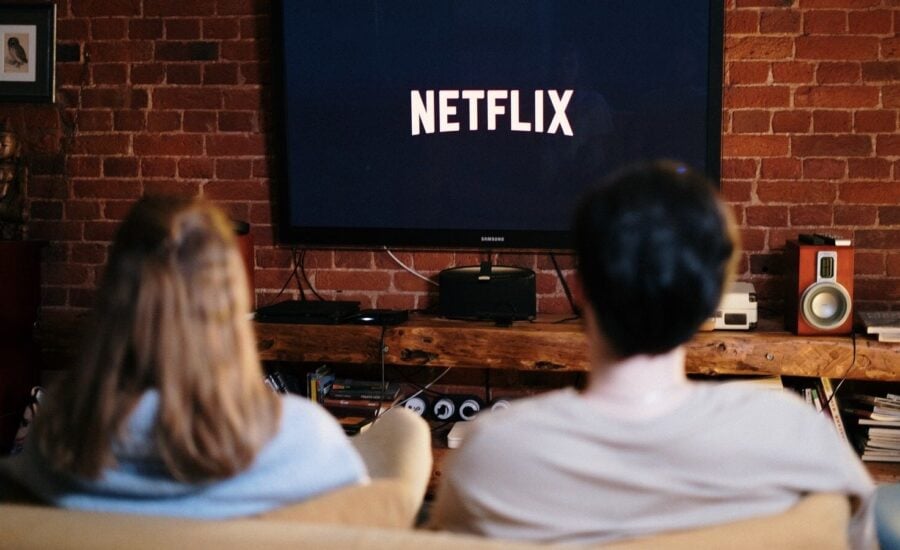 A couple is seen sitting on a sofa looking at a TV with it displaying Netflix logo