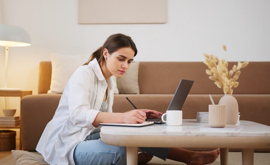 A woman sits at a coffee table and uses a laptop and a notepad.