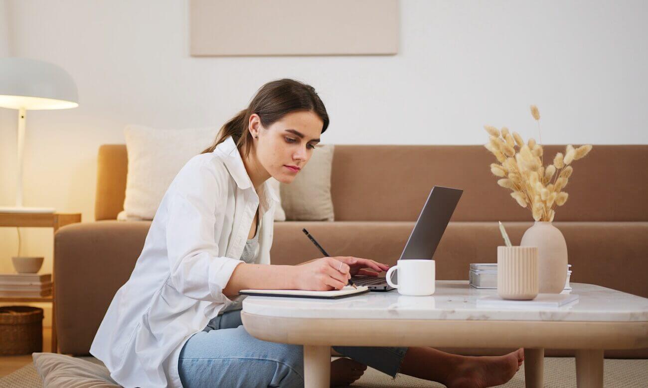 A woman sits at a coffee table and uses a laptop and a notepad.