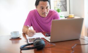 A man looks at his laptop and takes handwritten notes.