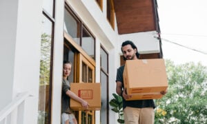 A man moving out from the family home, carrying a box of toys outside of the house,as his ex-wife walks behind him with another box.