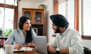 A couple sitting at their kitchen table, with a pad and paper in front of them as well as their computer.