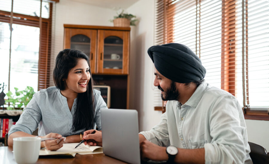 A couple sitting at their kitchen table, with a pad and paper in front of them as well as their computer.