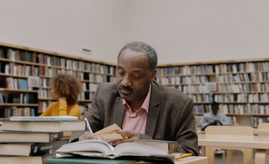 A man in his 50s, sitting in a college or university library, reading a book.