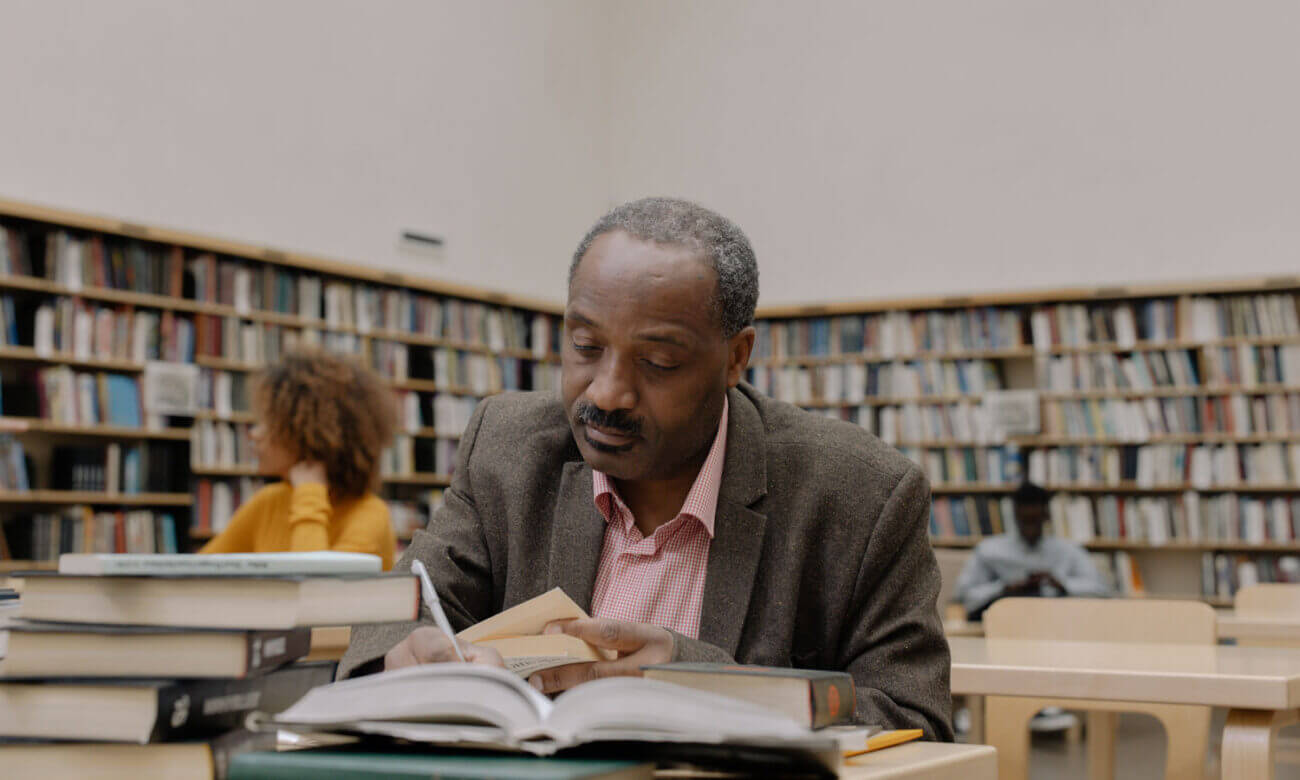 A man in his 50s, sitting in a college or university library, reading a book.
