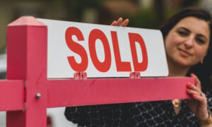 A woman in the background is adding a "sold" card to a "for sale" real estate sign.