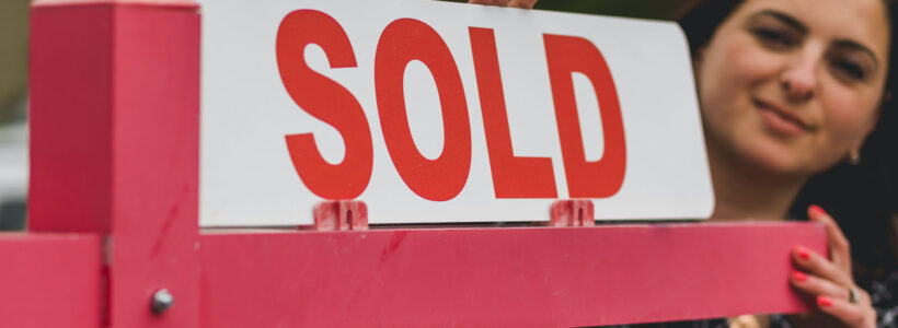 A woman in the background is adding a "sold" card to a "for sale" real estate sign.