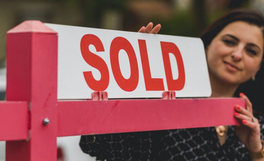 A woman in the background is adding a "sold" card to a "for sale" real estate sign.