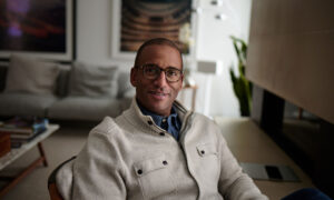 Duane Ledgister, wearing glasses and a thick cardigan over a button-up shirt, sits at his desk smiling.