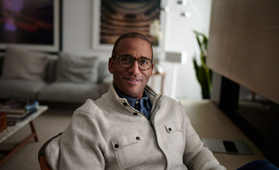 Duane Ledgister, wearing glasses and a thick cardigan over a button-up shirt, sits at his desk smiling.