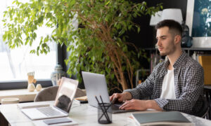 A young man sits a table with his laptop open, with an empty seat in front of him.