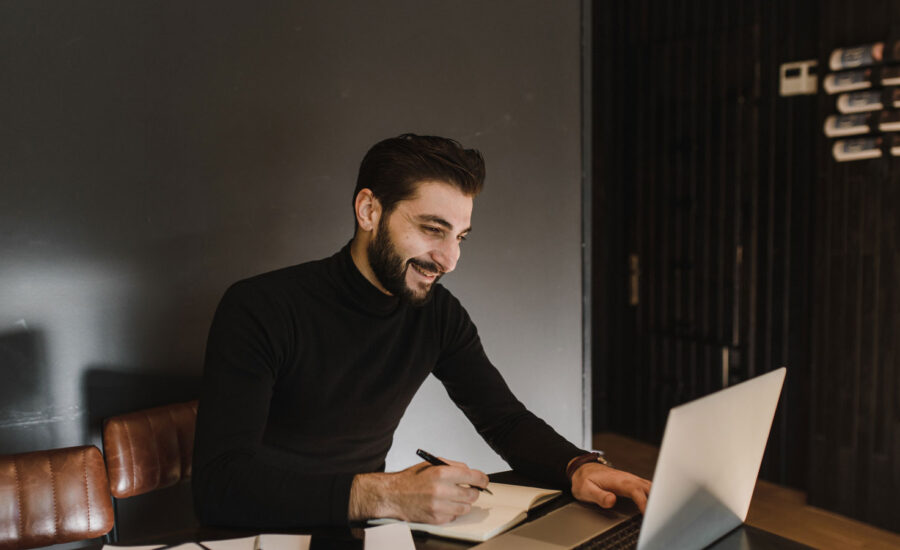 Man is smiling at his computer, as he takes notes of the money he would like to save on his new investing strategy