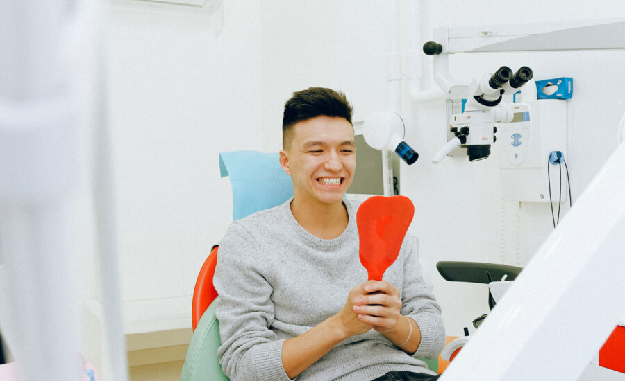 A man sits in a dentist chair, happy with the results of his latest check up, as holds a mirror inspecting his teeth.