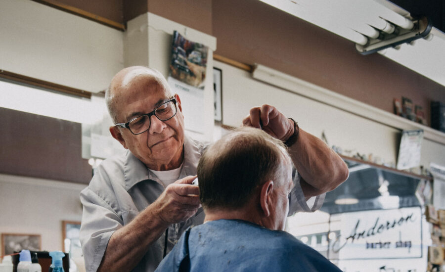 A man nearing the age of retirement is working as a barber, cutting the sideburns of another man in his barber chair.