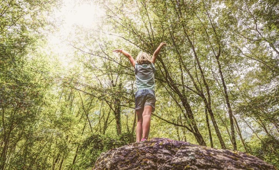 A young child in a forest, throwing their arms in the air