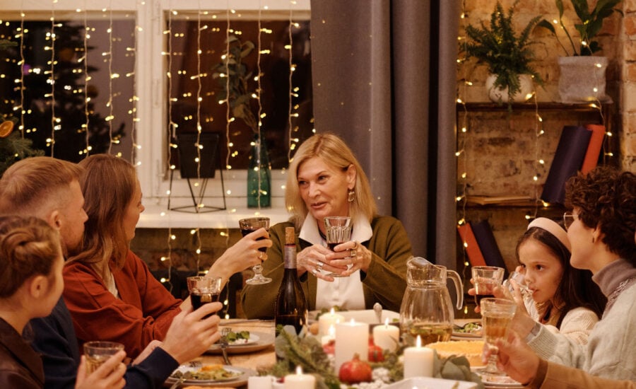 A woman in her early 50s sits at the head of a dinner table raising a glass to friends and family.