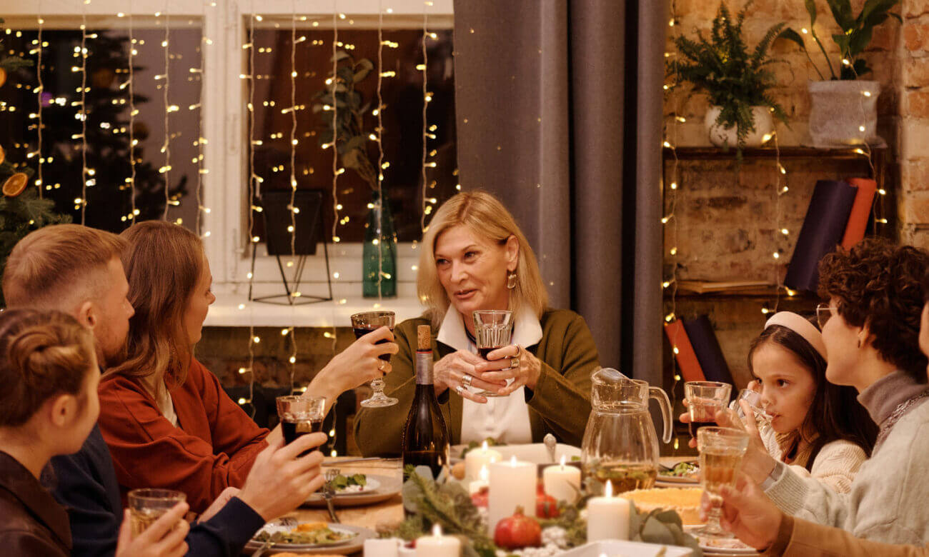 A woman in her early 50s sits at the head of a dinner table raising a glass to friends and family.