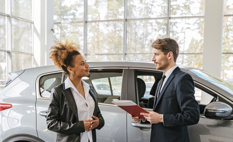 A woman is standing inside a auto dealership, talking with a man holding a folder, as he tells her the features about the car she is planning to buy. Negotiations are coming.