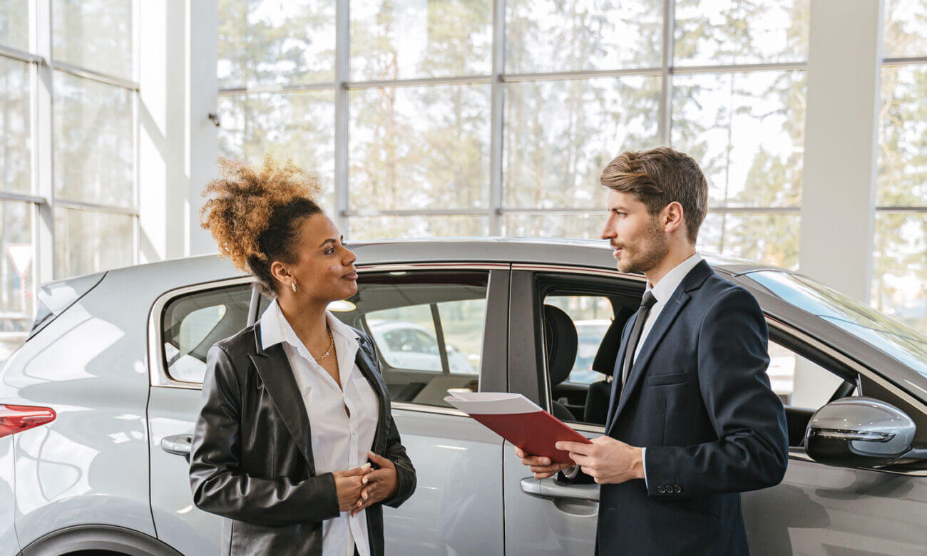 A woman is standing inside a auto dealership, talking with a man holding a folder, as he tells her the features about the car she is planning to buy. Negotiations are coming.