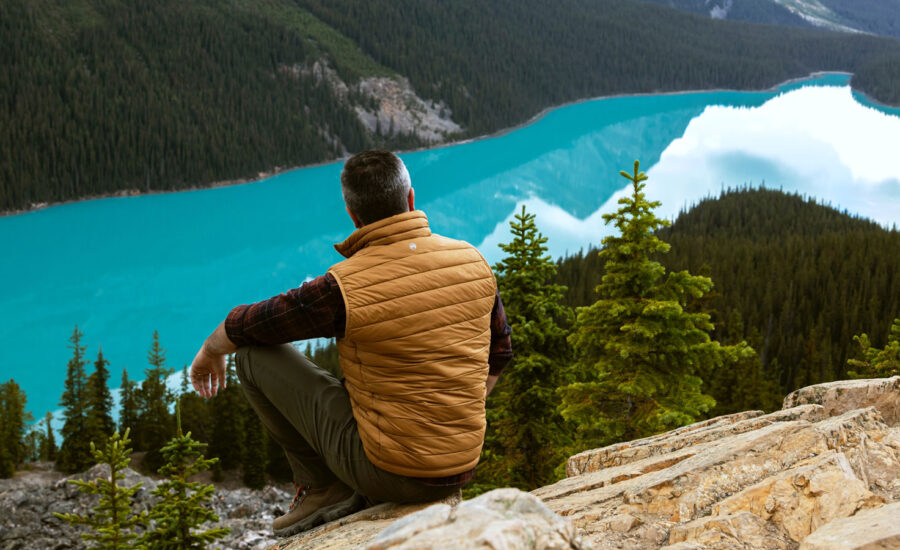 A man on a hike is sitting on top of a hill, looking out to a lake and mountains in the distance.