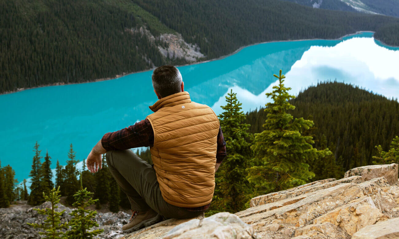 A man on a hike is sitting on top of a hill, looking out to a lake and mountains in the distance.