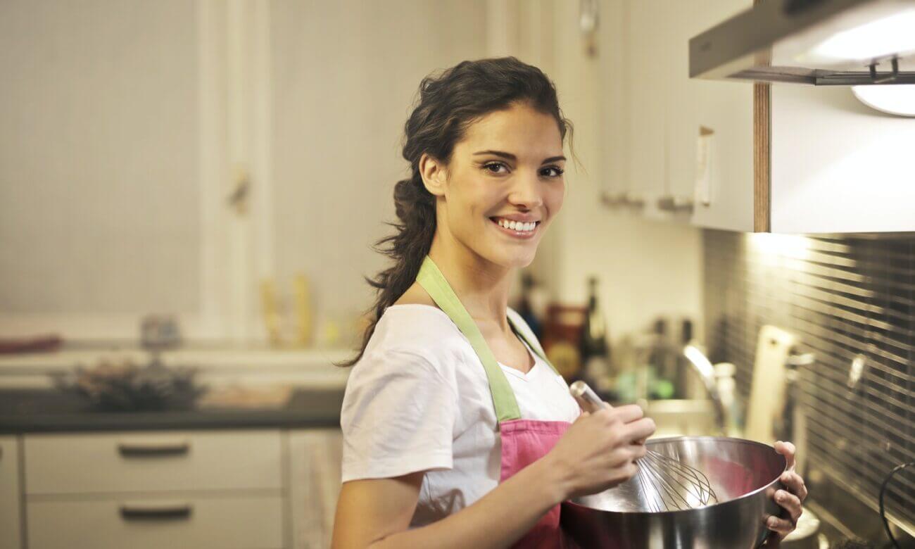 A smiling woman holds a mixing bowl and whisk in a kitchen