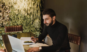 A man sits at his desk, making notes on paper as he reads about his investment accounts online