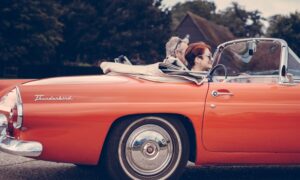 An older couple is seen riding in a car down a road in a classic car as they contemplate investing in classic car companies