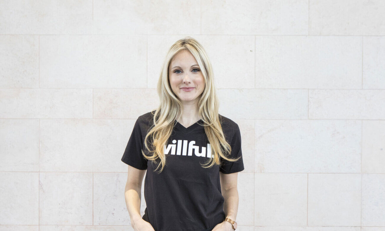 Erin Bury stands with her hands in her pockets as she leans against a tiled wall. She wears a T-shirt that reads Willful, her company's logo.