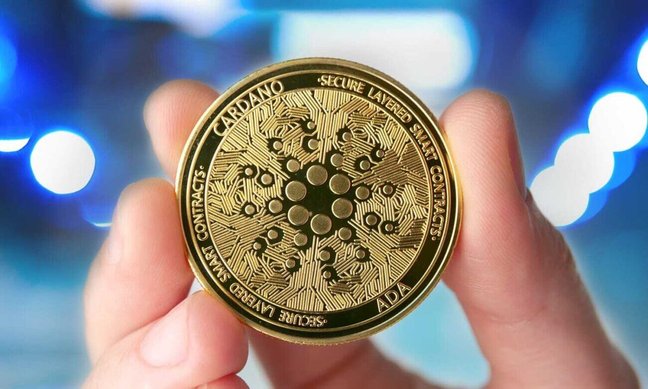 A hand holds up a gold coin with the Cardano logo