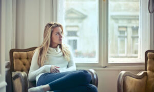 A woman is sitting on her couch in her apartment with a pensive expression on her face.